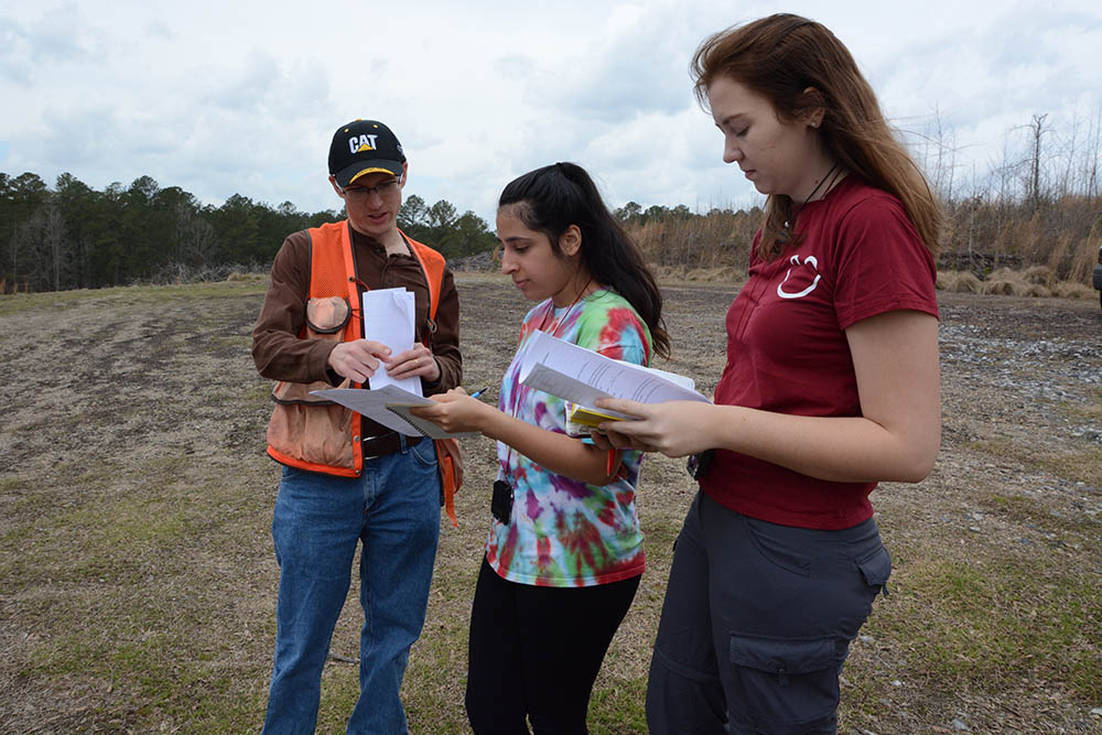 Two female students and their professor study their notes in the forest